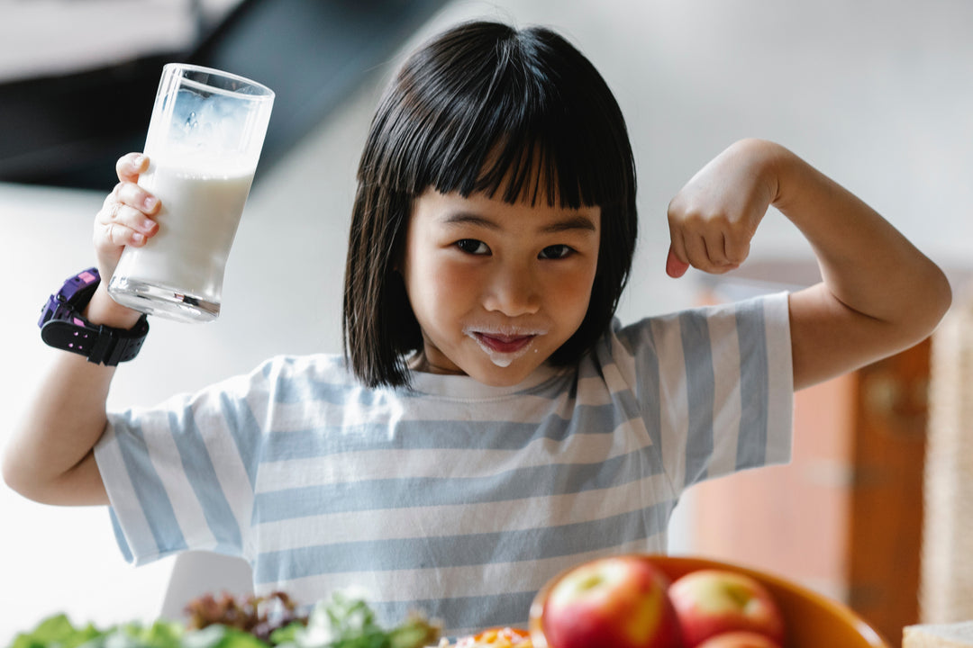 Jeune fille montrant ses muscles tout en tenant un verre de lait, avec des fruits et légumes en arrière-plan, illustrant la santé et l'énergie grâce à une bonne alimentation