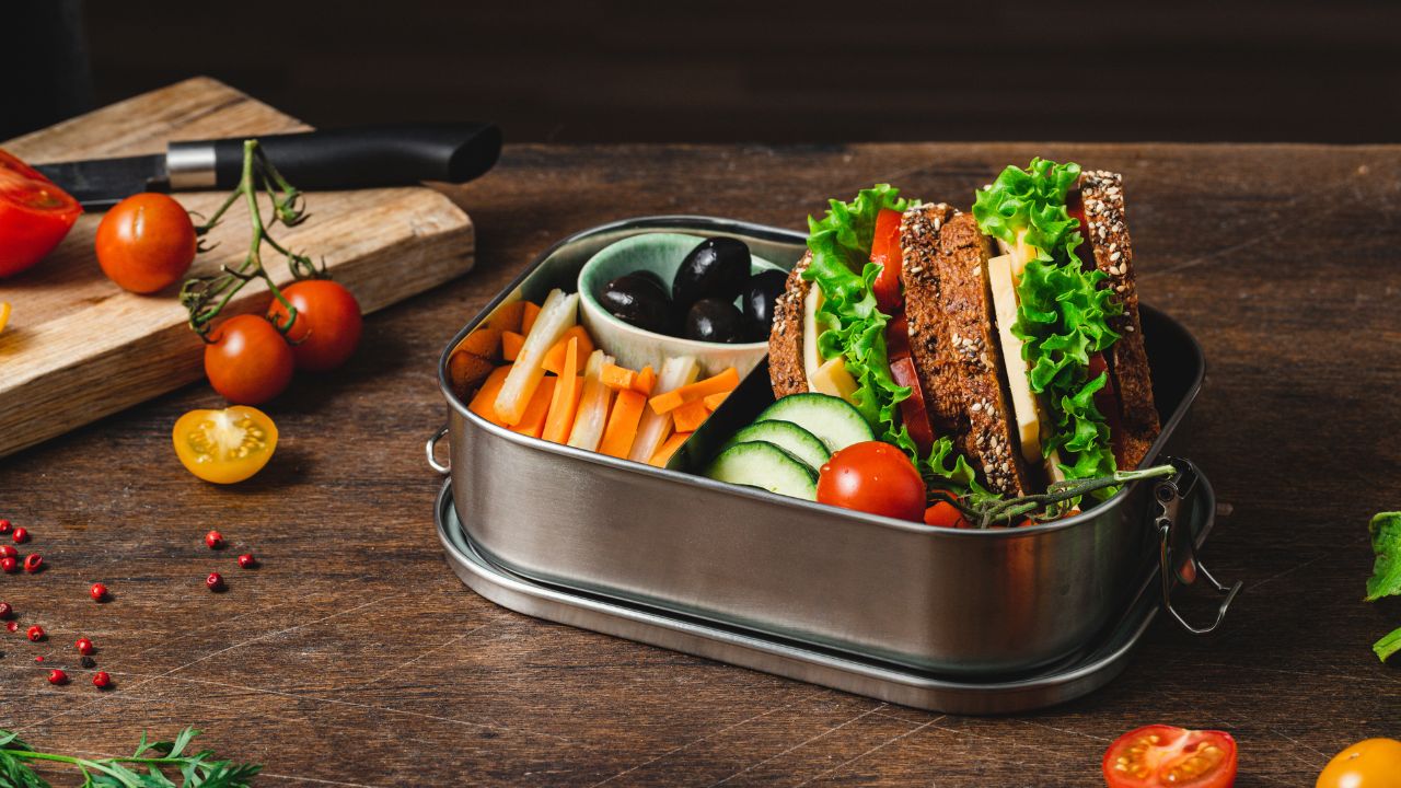 Lunch box en acier inoxydable contenant un sandwich végétarien avec laitue, tomate et fromage, des bâtonnets de carotte et de concombre, et des olives noires, posée sur une table en bois