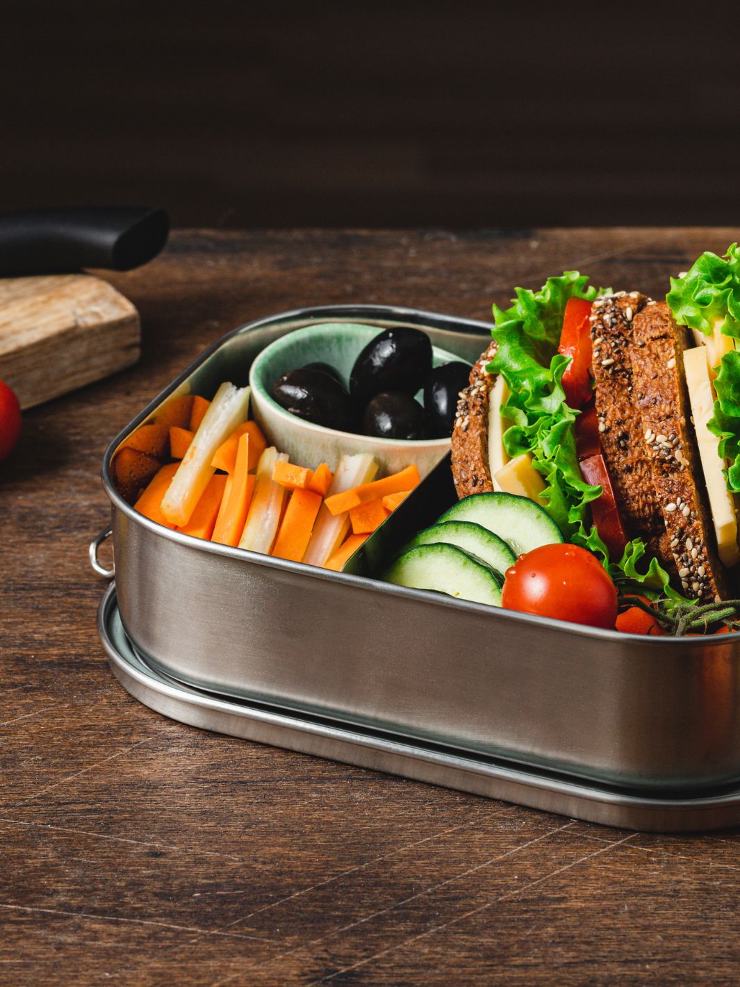 Lunch box en acier inoxydable contenant un sandwich végétarien avec laitue, tomate et fromage, des bâtonnets de carotte et de concombre, et des olives noires, posée sur une table en bois