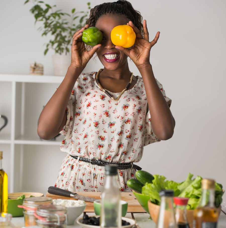 Femme souriante tenant un poivron vert et un poivron jaune devant ses yeux dans une cuisine, entourée d'ingrédients frais comme des légumes, de l'huile d'olive et des épices, préparant un repas sain